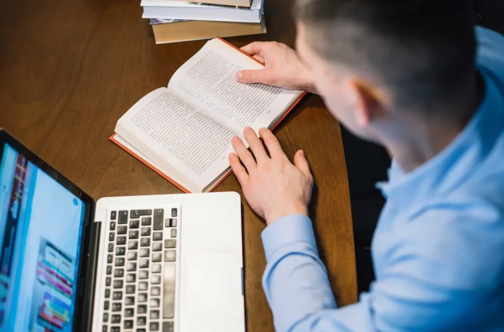 Man reading a book beside a laptop
