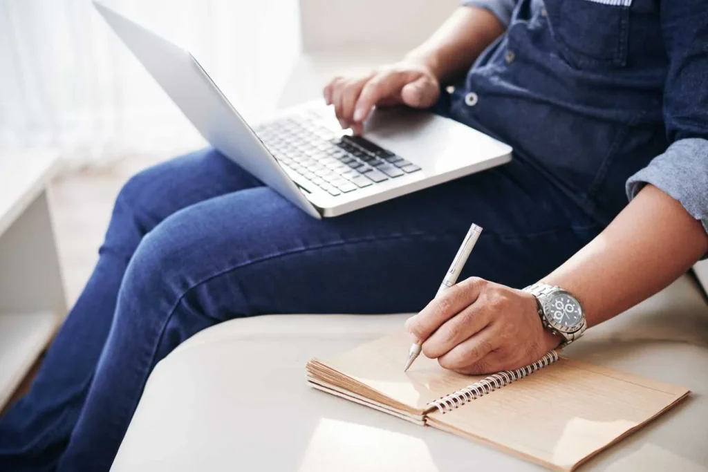 Person using a laptop while writing notes in a spiral notebook.