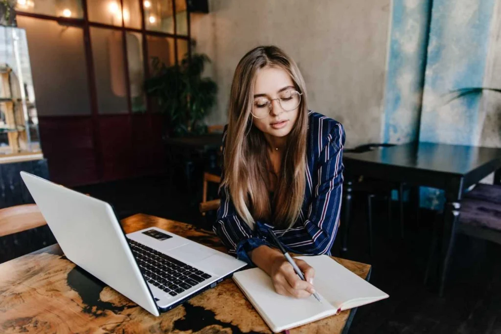Person writing in a notebook while working on a laptop in a caf&eacute;.