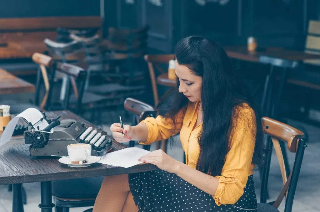 Person writing on paper beside a typewriter and a cup of coffee.