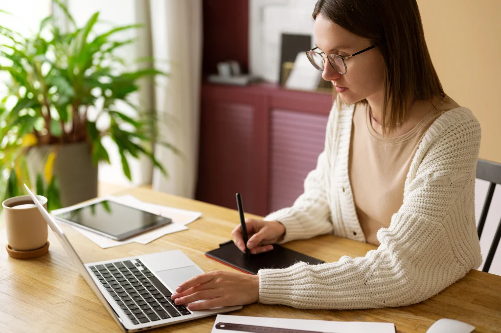 A woman with long hair sits at a dark desk, typing on a laptop with an open book nearby in a room with blue walls and bookshelves.