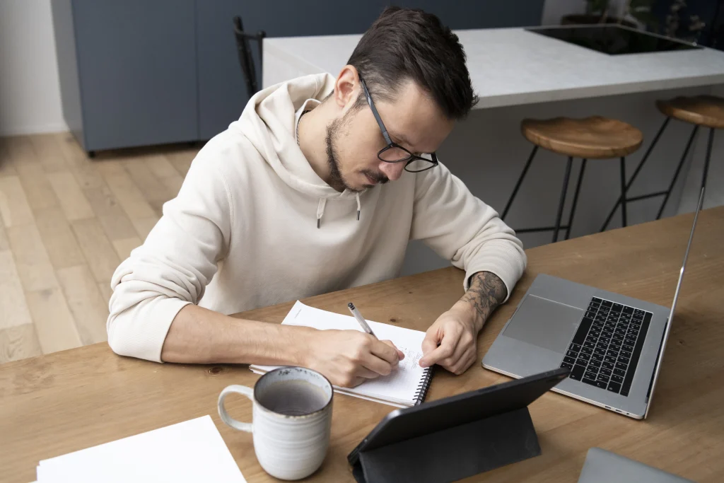 A man in a beige hoodie sits at a wooden desk, focused on writing in a notebook next to a laptop and coffee mug.